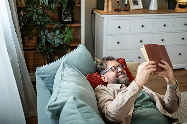 istock Mid adult man lying on sofa reading a book and smiling 2215393405