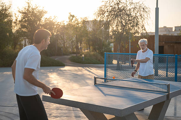 Senior father and son are engaged in an intense game of table tennis at a park during sunset. Shadows and light create dynamic patterns on the ground, adding drama to the scene.