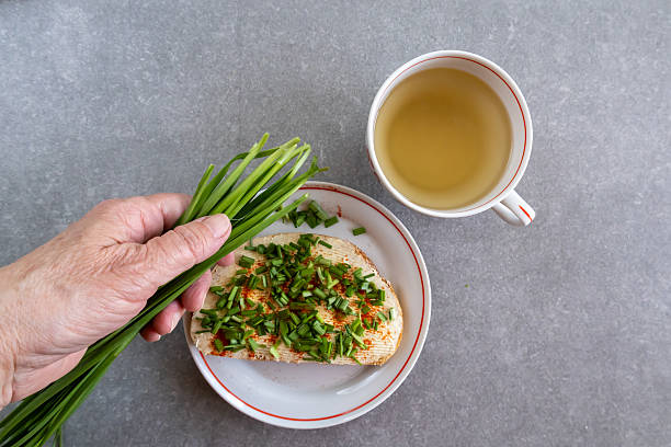 A hand holds a bunch of fresh chives beside a red striped plate with buttered bread topped with chives and paprika. A cup of tea completes the cozy scene on the gray countertop.