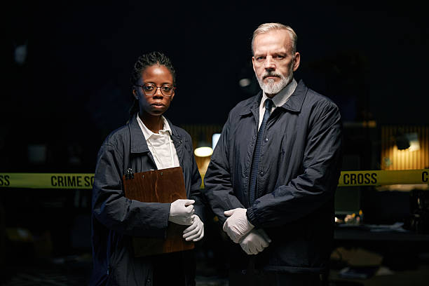 Multiracial detectives standing side by side, holding clipboard and wearing gloves in dimly lit room with crime scene tape in background. Portrait of two people looking straight at camera