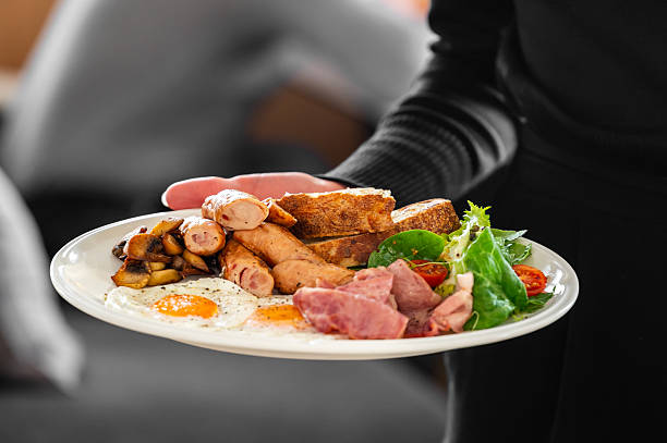 istock A plate with a traditional English breakfast, including sausages, fried eggs, mushrooms, bacon, toast, and a fresh side salad with cherry tomatoes, held by a person wearing a black sweater 2215414089