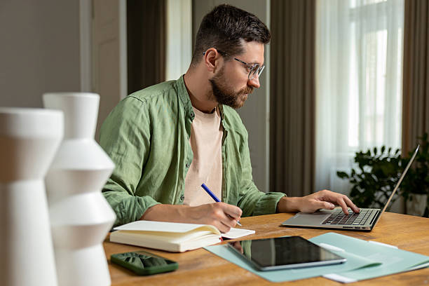 istock Bearded man reviewing notes on desk while working online with laptop and papers 2215417631