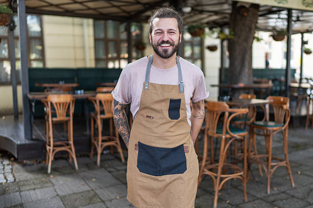 Portrait of a young, bearded waiter smiling confidently while wearing an apron, standing in front of his restaurant with hands casually tucked into pockets, embodying pride and happiness