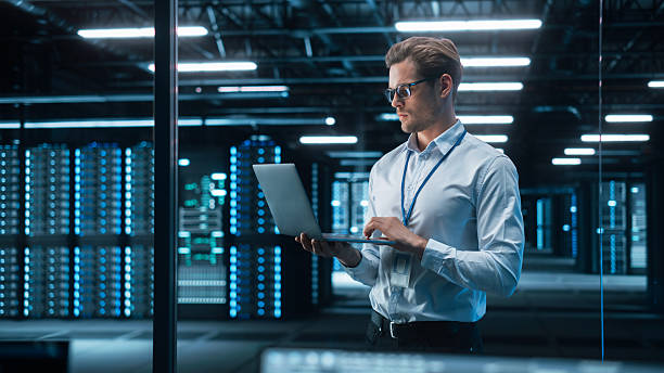 istock Portrait of IT Specialist Uses Laptop in Data Center. Server Farm Cloud Computing Facility with Male Maintenance Administrator Working. Cyber Security and Network Protection 2215422889