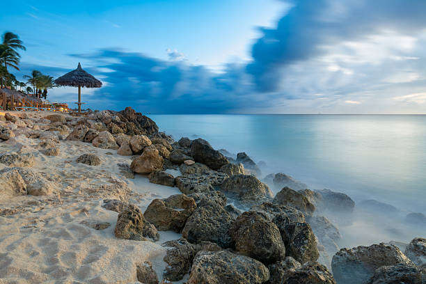Tranquil coastline at twilight in Aruba, features motion-blurred waves washing over rocks beneath a blue and cloudy sky, with a beach palapa.