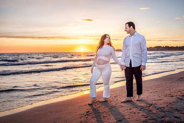 Happy pregnant couple walking hand in hand along the beach at sunset, savoring a romantic moment together while feeling the soft sand beneath their bare feet