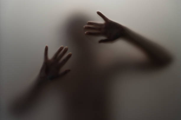 A hazy silhouette of a person is seen pressing their hands against a misted glass surface. The image evokes feelings of isolation, intrigue, and artistic abstraction through its moody lighting and obscured figure.