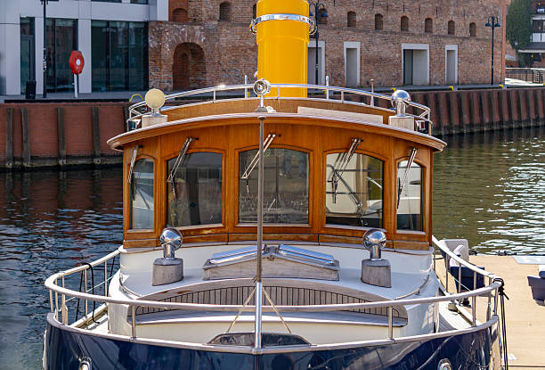 classic motorboat moored at a dock, featuring a wooden cabin structure and a distinctive yellow smokestack. In the background, a mix of red brick and glass urban-industrial architecture is reflected in the calm harbor water. This image is ideal for illustrating themes related to boating, water recreation, urban waterfront infrastructure, and nautical tourism.