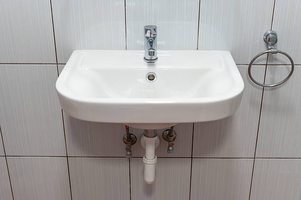 A close-up image of a modern ceramic bathroom sink mounted on a tiled wall. The sink features a sleek chrome faucet and is complemented by a metal towel ring to the side.