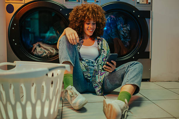 istock Young woman using smartphone while waiting for laundry at laundromat 2215453715