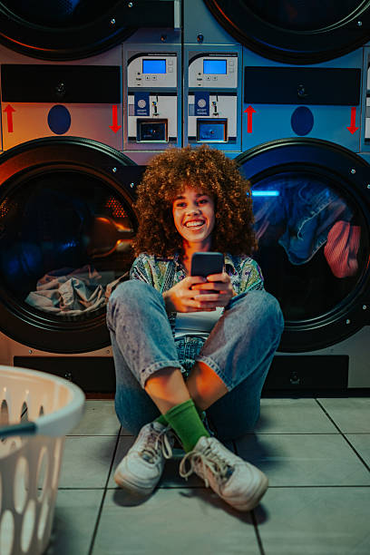 istock Young woman using smartphone while waiting for laundry at laundromat 2215453730