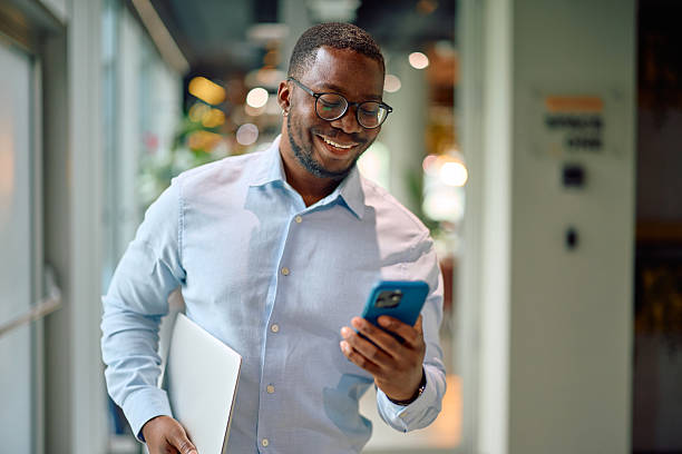 istock Smiling businessman walking in office corridor using smartphone and carrying laptop 2215454893
