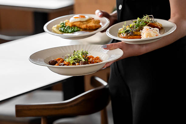 istock A restaurant server holds three plated meals, featuring pasta with meat and greens, a breaded cutlet with mashed potatoes and peas, and a dish with coleslaw. The setting suggests a dining experience 2215463831