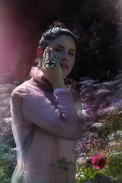 A young lady, dressed in a transparent top, holds an Delias Diaphana butterfly on her cheek, while standing in a Laceflower field in a color grading effect.