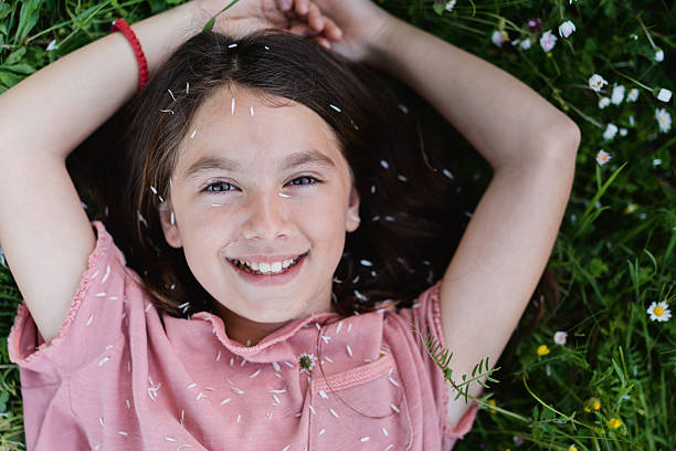 A young girl lying on the grass, looking up at the camera with a gentle smile. She has long brown hair and is wearing a pink shirt. White flower petals are scattered on her face, hair, and clothes, and there's a small daisy between her lips. The picture feels calm, natural, and playful.