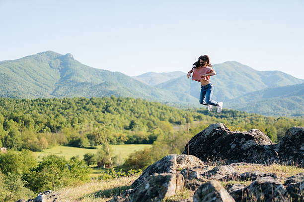 A young girl walking or jumping on large rocks in a scenic, mountainous landscape. She is wearing a pink top, blue jeans, and white sneakers. The background features rolling green hills and tall mountains under a clear blue sky. The image captures a feeling of freedom, adventure, and connection with nature.