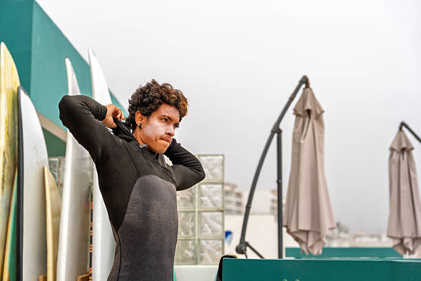 Young adult surfer putting on his wetsuit on the balcony of a beach house, preparing for a surf session