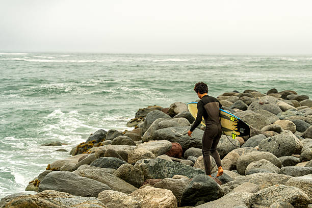 Young surfer carrying a surfboard, walking down rocky terrain toward the ocean waves under a cloudy sky, embracing an active lifestyle