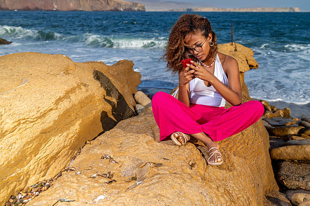 Young woman relaxing on a rock at the beach, engaging with her mobile phone while enjoying the beautiful ocean view under the summer sun