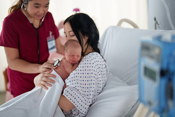 A female pediatric nurse of Asian decent makes rounds to her patients as she spends time in a new mothers room, checking her baby's vital signs.  The Infant is laying peacefully in a diaper on his mothers chest as the nurse listens closely with her stethscope.