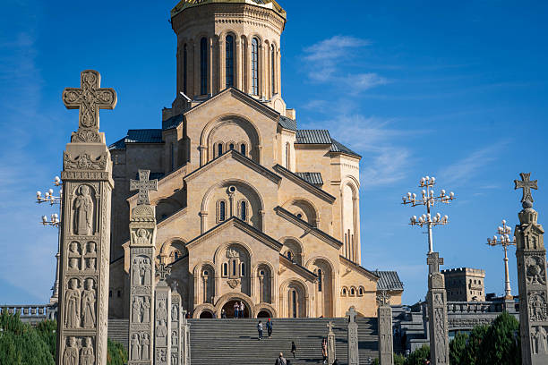 Tbilisi, Georgia - October 29, 2024: A view of the majestic Holy Trinity Cathedral of Tbilisi (Sameba) under a clear blue sky, framed by intricately carved stone pillars and crosses in the foreground. People can be seen ascending the grand staircase leading to the cathedral, highlighting the scale and importance of this religious landmark in Georgia. The warm tones of the cathedral's facade contrast with the sky, emphasizing its architectural grandeur and spiritual significance