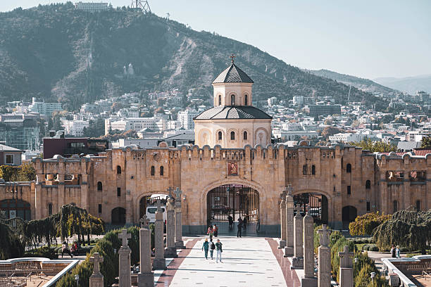 Tbilisi, Georgia - October 30, 2024:  A wide shot captures the Svetitskhoveli Cathedral complex in Mtskheta, Georgia, showcasing its impressive stone architecture, including arched gates, towers, and the main cathedral with its conical dome. People stroll along the paved pathways within the complex. In the background, the city of Mtskheta spreads out towards rolling hills under a bright sky. The scene highlights the historical and religious significance of this UNESCO World Heritage site