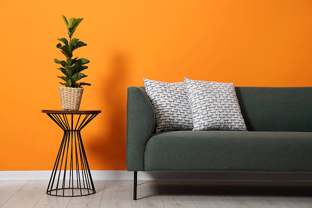 istock Stylish sofa with cushions and plant on side table near orange wall 2215514555
