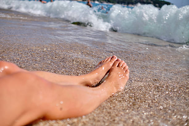 A woman relaxes on the beach with her bare feet stretched out on the sun-warmed sand, as soft ocean waves gently approach. This serene moment captures the peaceful essence of a summer escape, evoking tranquility, freedom, and connection with nature.