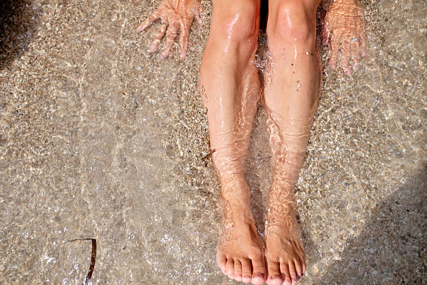 A woman relaxes on the beach with her bare feet stretched out on the sun-warmed sand, as soft ocean waves gently approach. This serene moment captures the peaceful essence of a summer escape, evoking tranquility, freedom, and connection with nature.