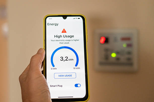 Hand holding smartphone displaying high electricity usage alert from smart plug application, with blurred electricity meter in background, promoting energy saving and cost reduction