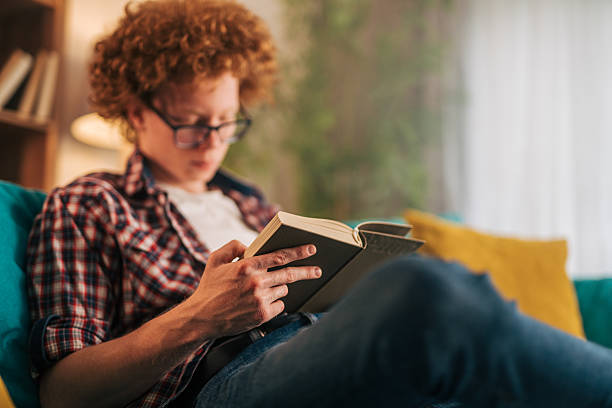 istock Young man reading a book on the sofa at home 2215522736