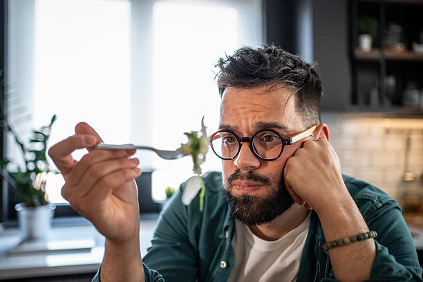 Sad young man disgusted by unhealthy food on fork in kitchen