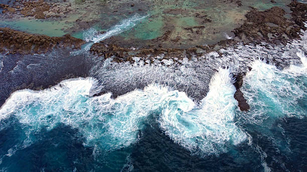 Waves Crashing Against Rocky Hawaiian Coastline. Top-down view of ocean waves forcefully crashing over jagged volcanic rocks and shallow tide pools along a Hawaiian shoreline.