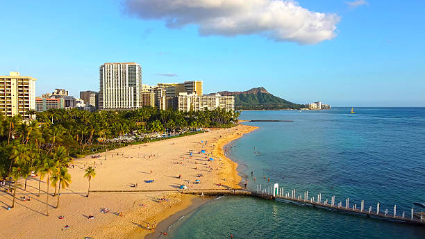 Waikiki Beach and Diamond Head at golden hour. Aerial view of Waikiki Beach with palm trees, people on the sandy shore, blue ocean waters, city buildings, and Diamond Head crater in the background under a sunny sky