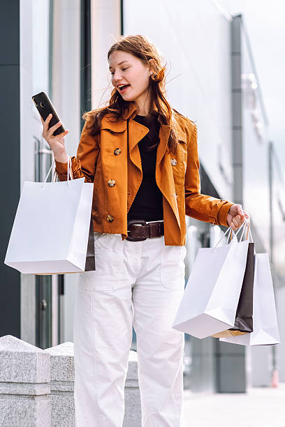 istock A young woman stands outside a contemporary building, checking her phone while holding shopping bags. She appears excited as she arranges for her taxi.Purchases, black friday, discounts, sale concept. 2215533644
