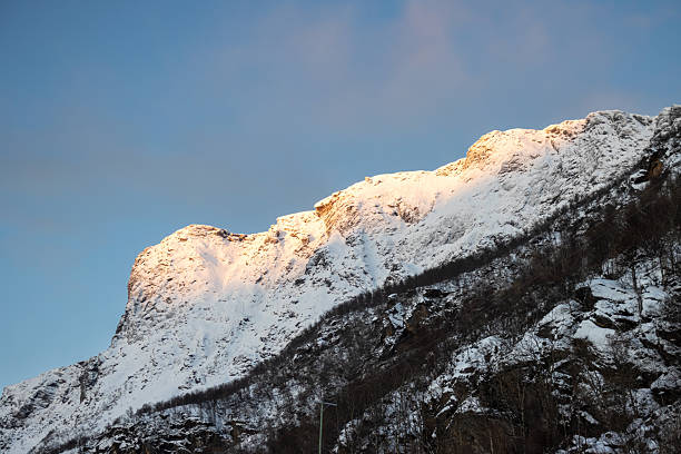 Detail of a mountain. Forest in the lower part and sharp edge of the moutain. All covered by snow in the arctic late autumn. Blue sky with light white clouds. Harstad, Norway.