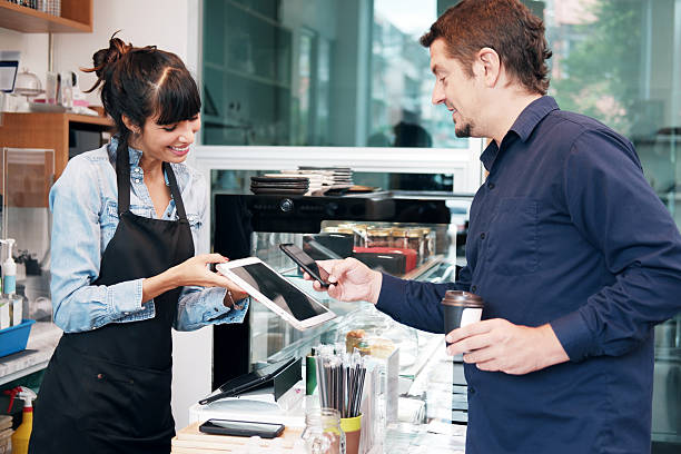 istock Young Caucasian barista woman is holding tablet for customer using smart phone scan QR code on tablet for payment at counter bar at coffee shop. Technology of digital pay without money with mobile banking application 2215554429
