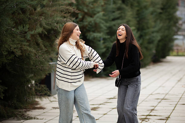Two young women share a joyous moment outside as they laugh and walk along a paved path surrounded by trees and nature.