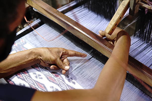 A detailed shot showcasing a person operating a traditional handloom to weave a patterned textile. Focus on the hands guiding the threads, the intricate details of the woven fabric, and the wooden structure of the loom. Capture of the crafting process, showcasing traditional textile production.
