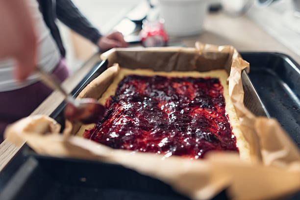 A rectangular tart cake rests on a parchment-lined baking tray as a young woman carefully spreads a layer of fruit jam on top using a spoon. The rich, glossy texture of the jam contrasts with the soft pastry base. In the background, a kitchen counter displays various baking essentials.\nShot with Canon R5