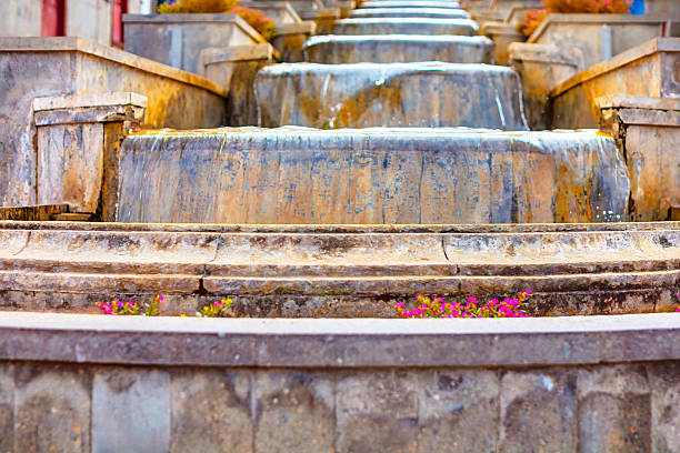 Weathered stone fountain with cascading water flows down multiple tiers, showing signs of age and moss. Pink flowers bloom along the base, adding a touch of color to the gray structure