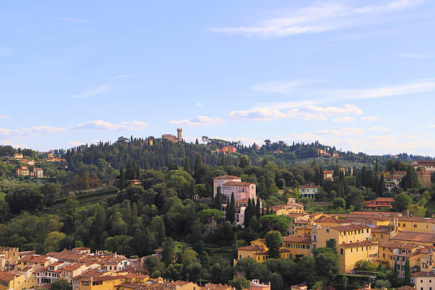 A scenic view of a hilly landscape in Florence, Tuscany, Italy. The foreground features historic buildings with terracotta roofs nestled among lush greenery. Cypress trees are scattered throughout the scene, typical of the Tuscan countryside. In the background, a hill is topped with a few notable structures, likely villas or small castles, adding to the picturesque setting. The sky is clear with a few clouds, enhancing the tranquil atmosphere.