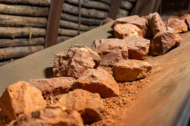 Chunks of reddish bauxite ore lie on a conveyor belt inside a dimly lit mining tunnel. The raw stones, with rough textures and earthy tones, are part of the aluminum production process.