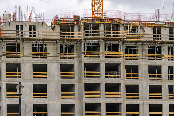 istock View of a multi-story building under construction, with visible window openings, formwork, and scaffolding. The concrete structure is in the raw state, and a portion of a tower crane is visible at the top. In the foreground, there's a classic street lamp. 2215608009