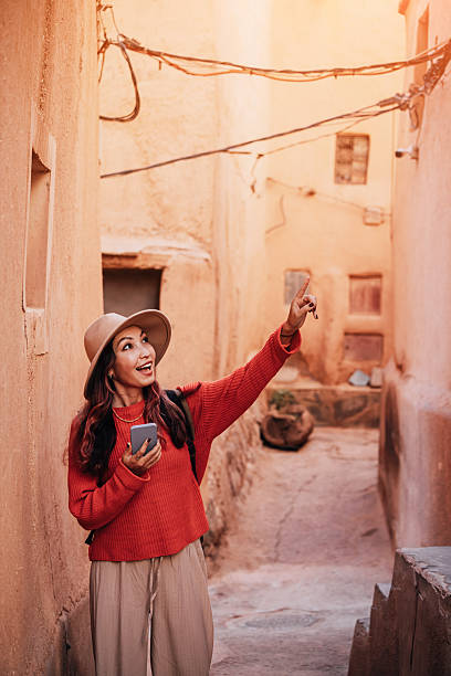 istock Young female tourist pointing at something interesting while exploring narrow alley in ancient Moroccan town, holding smartphone and wearing backpack 2215629363
