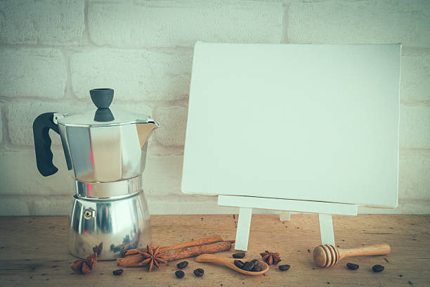 Wooden easel, white  blank canvas and coffee pot on wooden table with white wall background. Coffee shop, creative space and so on concept. Vintage tone.