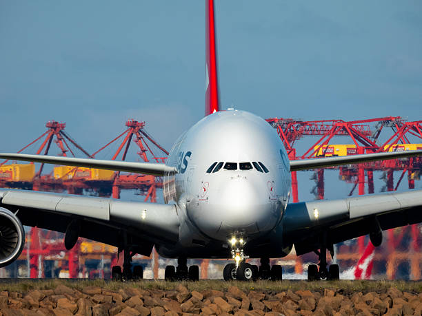 A Qantas Airbus A380-842 plane, registration VH-OQJ, taxiing to the international terminal after landing at Sydney Kingsford Smith Airport as flight QF64 from Johannesburg. The pilot and co-pilot are visible in the cockpit and wearing sunglasses. The wings remain in the landing configuration. \n\nIn the foreground are rocks forming the sea wall on Botany Bay. In the bottom right background is a Qantas Boeing B737-838 plane, registration VH-VXJ, taxiing from the third runway after arriving as flight QF740 from Adelaide. In the far background are cranes at the Port Botany Container Terminal. \n\nThis image was taken from near Kyeemagh Beach, Botany Bay on a cold, windy and sunny afternoon on 17 May 2025.