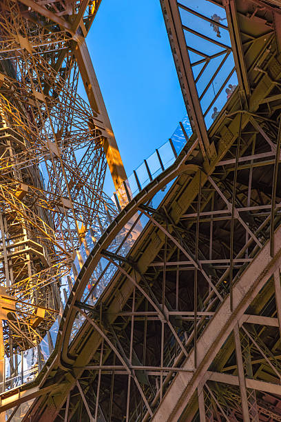 Interior structural view of the Eiffel Tower with people walking and taking photos on a glass-floored walkway.