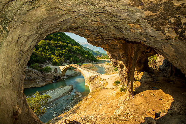 A limestone cave frames Ura e Kadiut (Kadiu Bridge) in Banje Permet, where a stone arch spans the turquoise Vjosa River in the lush Albanian Landscape