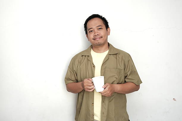 istock Calm Asian man holding a white mug, standing casually against a plain wall, dressed in a light green shirt and beige t-shirt, expressing relaxed morning vibes. 2215698970